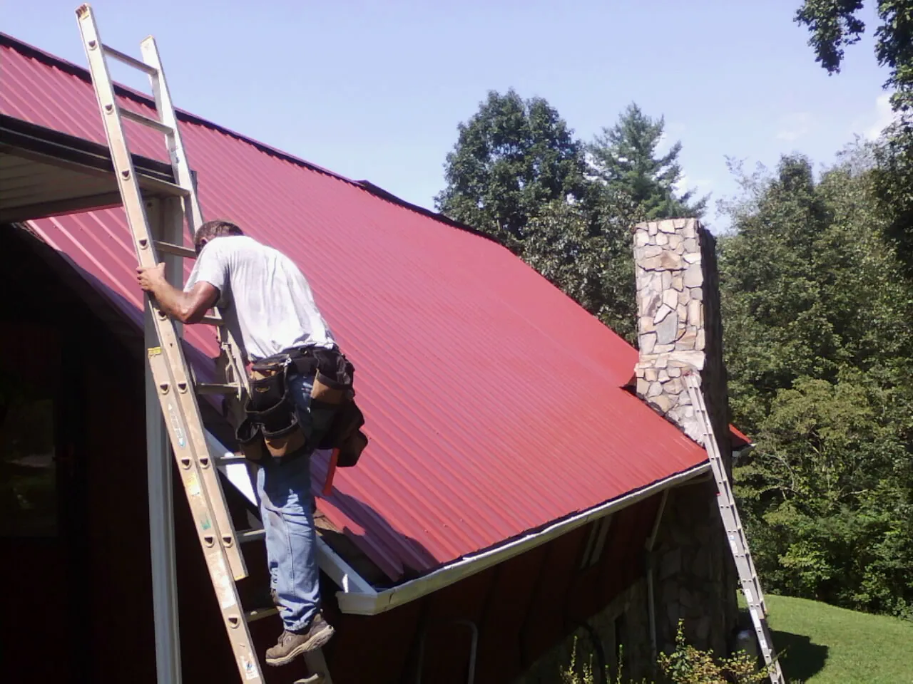 Roofer on a ladder beside a red metal roof and stone chimney — Soffit & Fascia Repair by Roofing Brothers in Pennsville